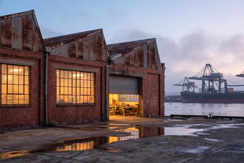 The brick factory with its roller door half open and lights on at dawn, harbour cranes behind