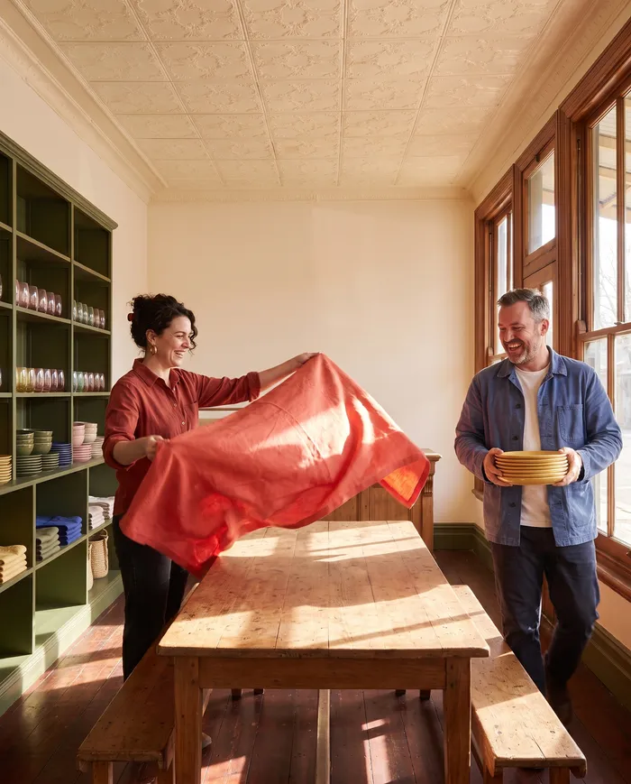 Lucia Ferraro shaking out a tomato red tablecloth over the long table in the shop while Sam Whitlock carries a stack of butter yellow plates