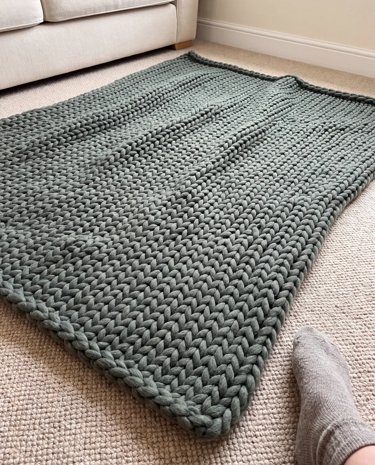 A green chunky knit throw laid flat on beige carpet in a bare living room, photographed from above on a phone with the photographer’s foot in shot.