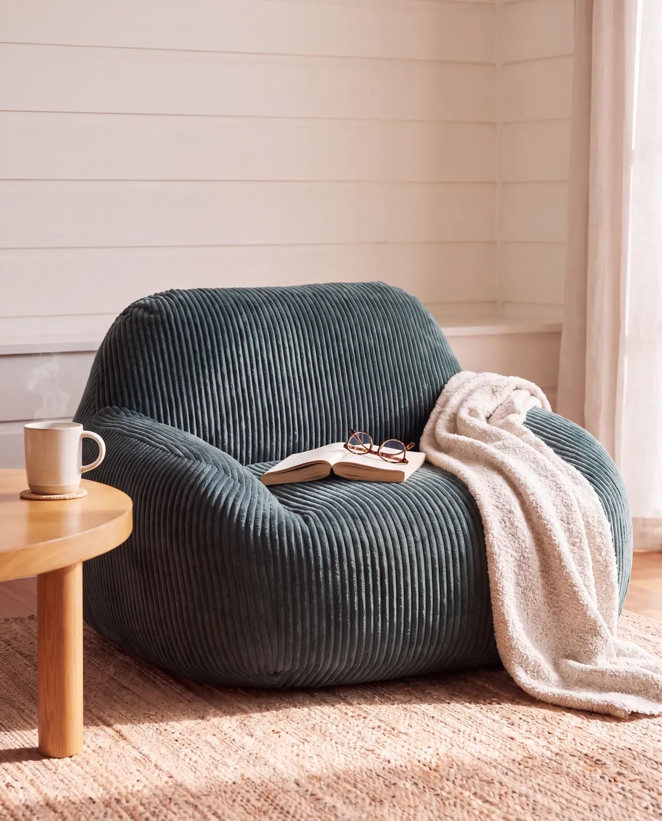 A green corduroy armchair with a cream throw, an open book on the seat and a mug on the side table.